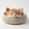 An orange and white cat sleeping comfortably inside a beige chunky knit cat bed with ear-like protrusions, set against a clean white background.