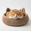 An orange and white cat sleeping comfortably inside a round, chunky knit cat bed with pointed ears, set against a clean white background.