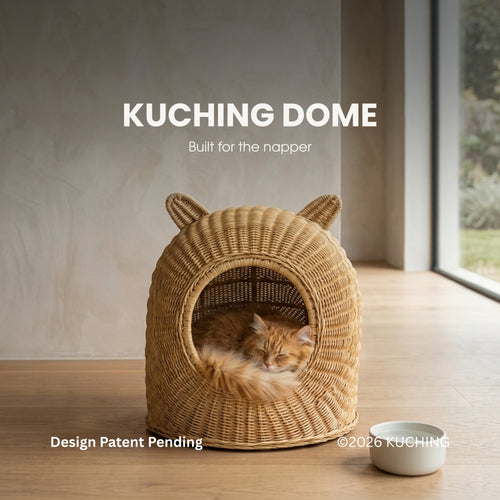 A wicker-style cat bed with ear-like protrusions sits on a wooden floor in a softly lit room, with an orange tabby cat curled up inside. White text at the top reads 'KUCHING DOME' and 'Built for the napper'; a small white bowl is placed nearby.