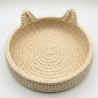 A round, hand-crocheted beige basket with cat ears on the rim, photographed on a clean white background.