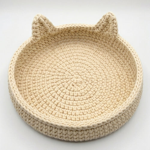 A round, hand-crocheted beige basket with cat ears on the rim, photographed on a clean white background.