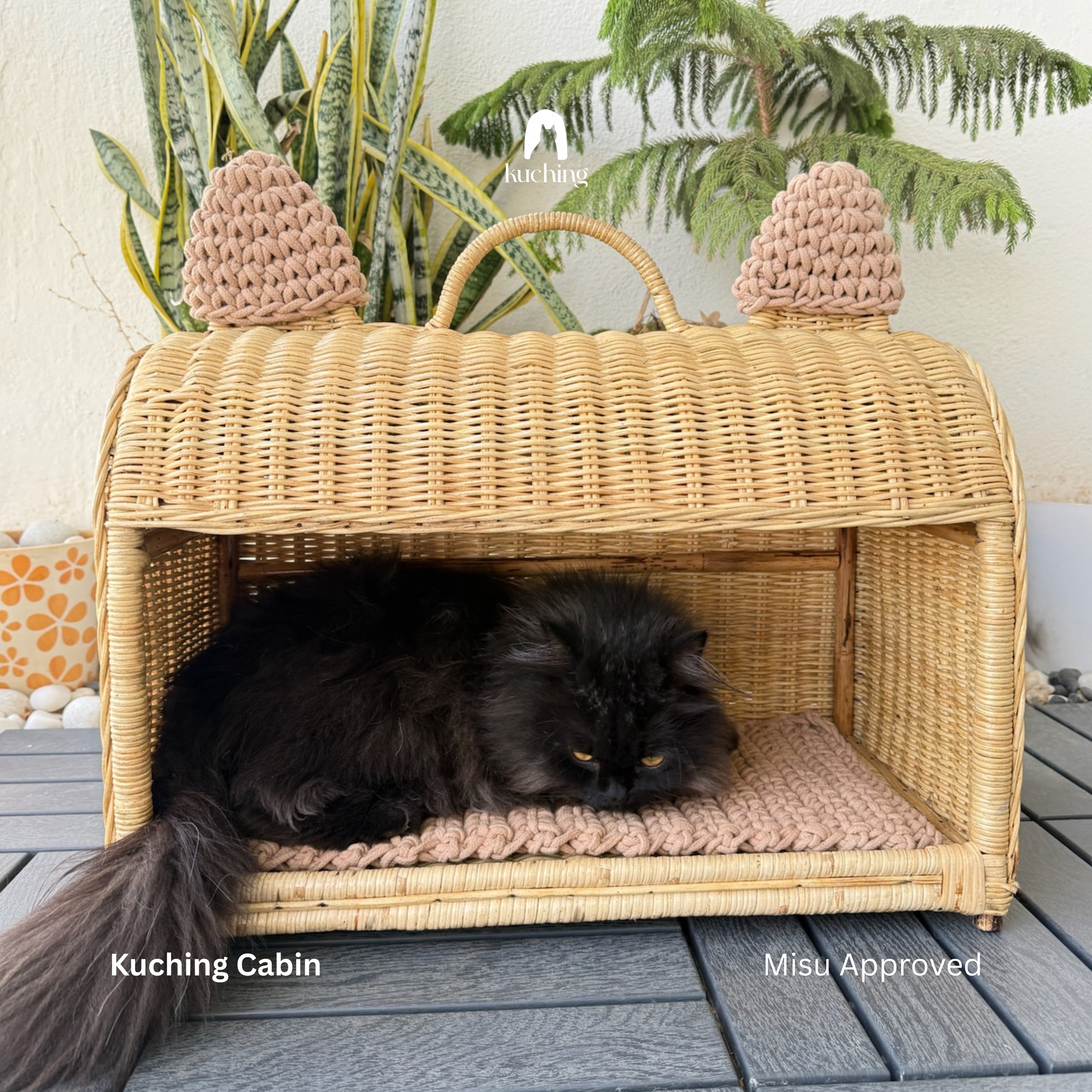 A fluffy black cat rests inside a woven wicker kigurumi cat house with knit ear covers and a cushioned floor, placed on a gray deck with plants in the background. Text overlays read 'Kuching Cabin' and 'Misu Approved'.
