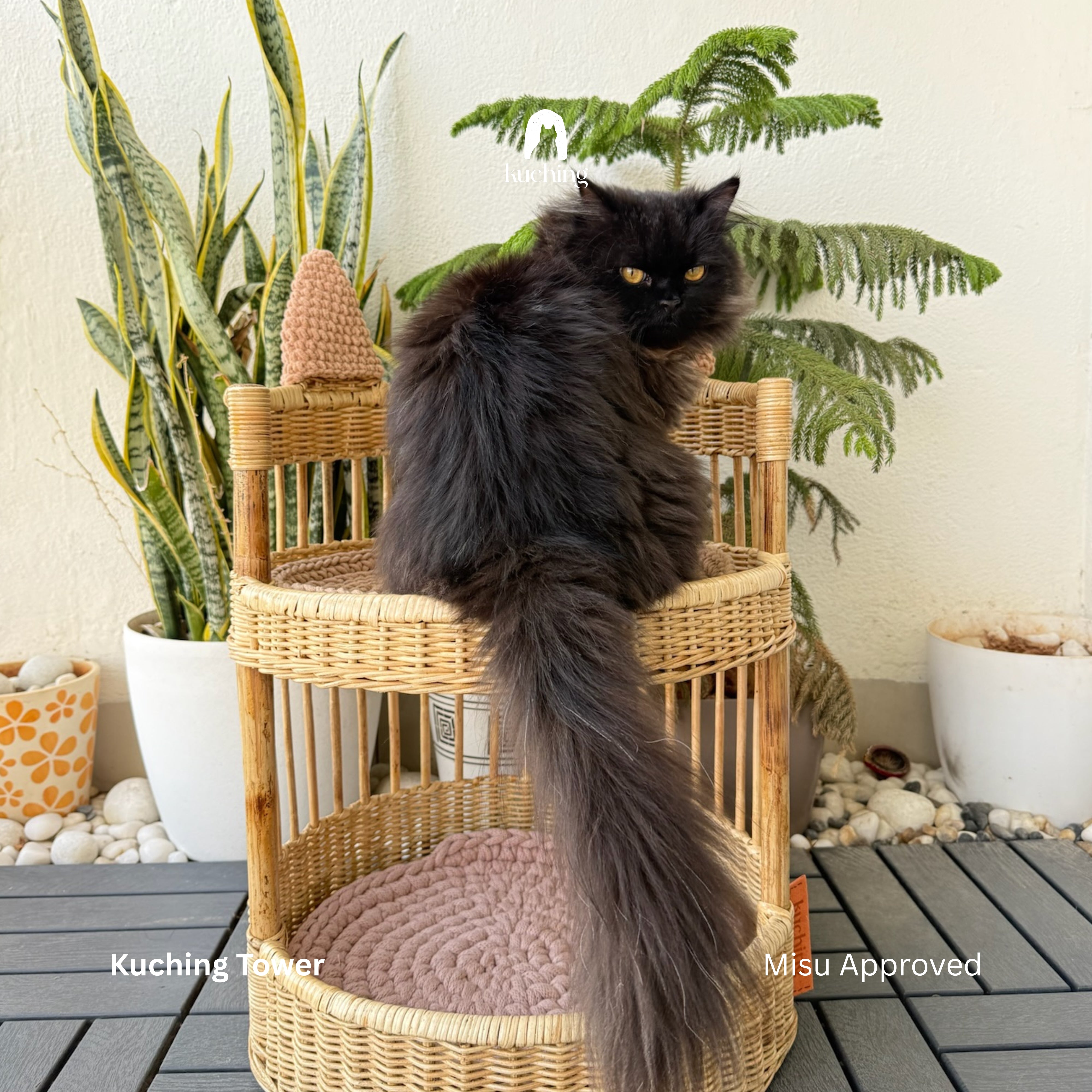 A fluffy black cat sits on a round woven rattan cat tower with a pink cushion, set on a dark gray deck with potted plants in the background. Text overlays read 'Kuching Tower' and 'Misu Approved'.