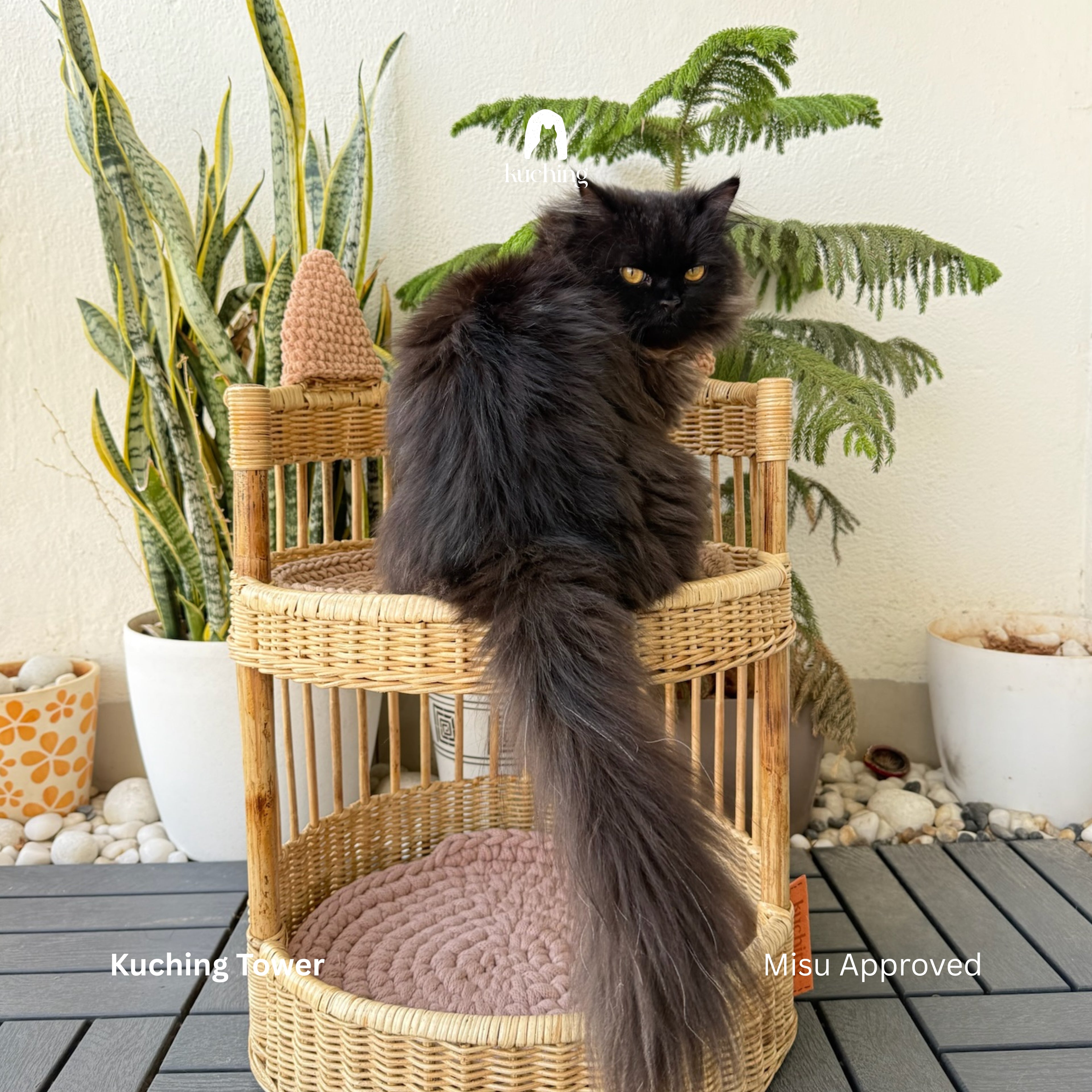 A fluffy black cat sits on a round woven rattan cat tower with a pink cushion, set on a dark gray deck with potted plants in the background. Text overlays read 'Kuching Tower' and 'Misu Approved'.